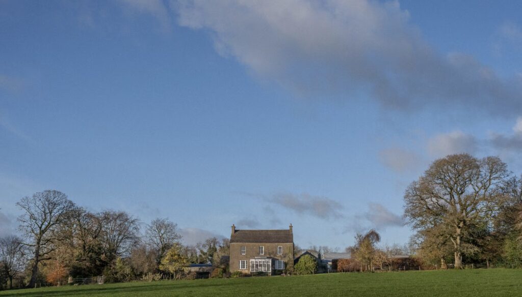 Stone country house centered beyond a grassy field, with bare trees on both sides under a blue sky with scattered clouds.